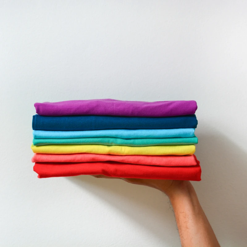 A hand holding a stack of neatly folded, plain, multi-colour T-shirts, ideal for customisation into branded T-shirts.